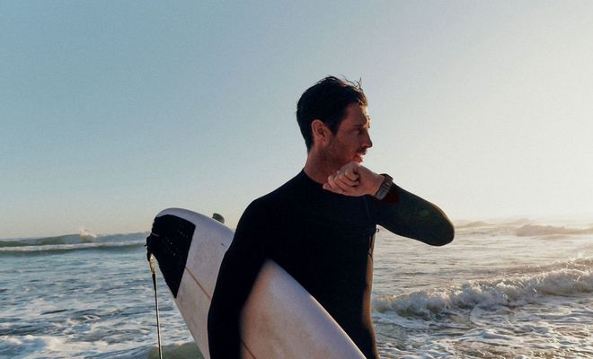 Surfista con traje de neopreno consultando su reloj inteligente fenix 9 mientras sujeta una tabla de surf en la orilla del mar.