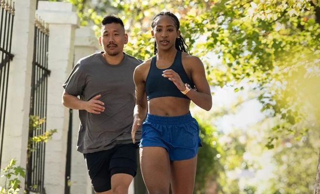 Pareja practicando running en una zona urbana con árboles y vallas de forja, la mujer luce un reloj inteligente en su muñeca izquierda.