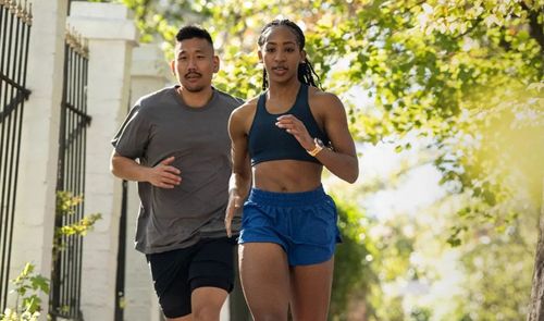 Pareja practicando running en una zona urbana con árboles y vallas de forja, la mujer luce un reloj inteligente en su muñeca izquierda.