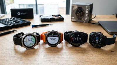 Cuatro relojes inteligentes Garmin Fenix alineados sobre una mesa de madera junto a herramientas de precisión y su caja original.