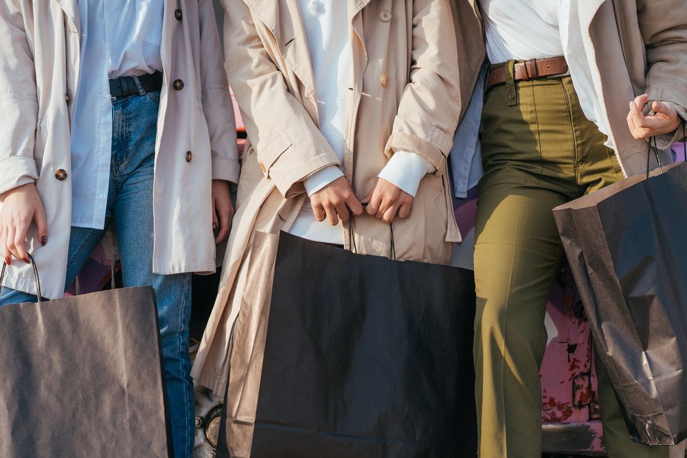 Tres personas con gabardinas beige sujetan bolsas de papel negras tras realizar compras.