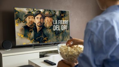 Persona sentada frente a un televisor viendo el programa La fiebre del oro de Discovery Channel mientras sostiene un bol de palomitas.