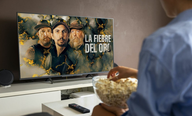 Persona sentada frente a un televisor viendo el programa La fiebre del oro de Discovery Channel mientras sostiene un bol de palomitas.