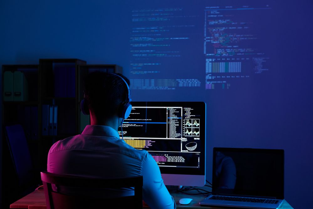 Persona trabajando de noche frente a un monitor con líneas de código y un proyector de fondo.