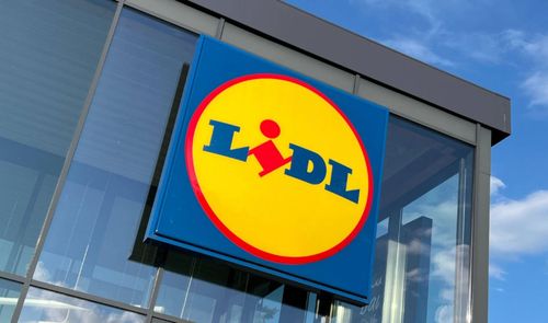 Logotipo circular amarillo y azul de Lidl instalado en una fachada de cristal bajo un cielo despejado.
