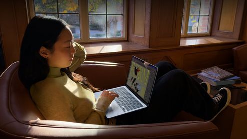 Mujer joven sentada en un sillón utilizando un ordenador portátil MacBook Air en una sala iluminada por luz natural.