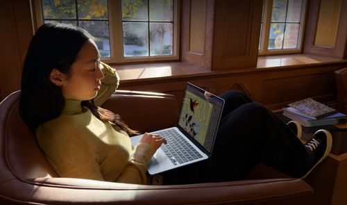 Mujer joven sentada en un sillón utilizando un ordenador portátil MacBook Air en una sala iluminada por luz natural.