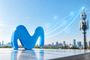 Logotipo de Movistar en primer plano frente a una torre de telecomunicaciones con iconos de conectividad sobre una ciudad.