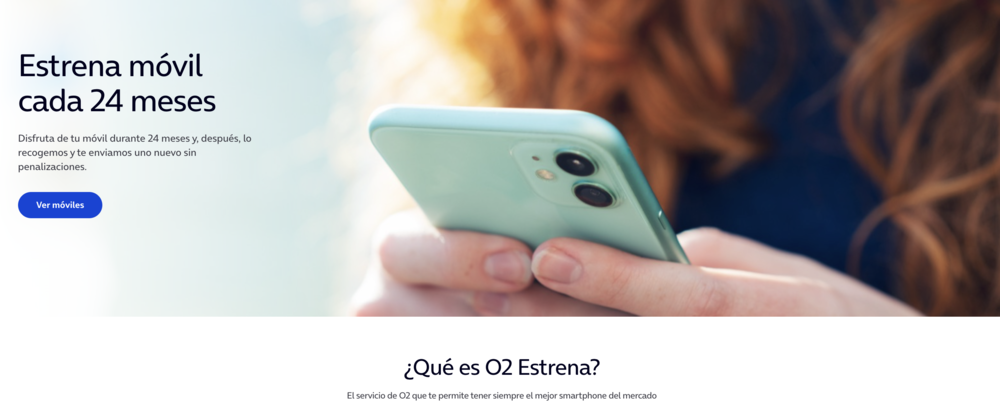 Primer plano de una persona sosteniendo un teléfono móvil verde claro con doble cámara trasera frente a un banner publicitario del servicio O2 Estrena.