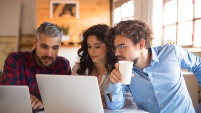 Tres personas jóvenes observando con atención la pantalla de un ordenador portátil en un entorno de oficina o cafetería.