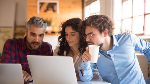 Tres personas jóvenes observando con atención la pantalla de un ordenador portátil en un entorno de oficina o cafetería.