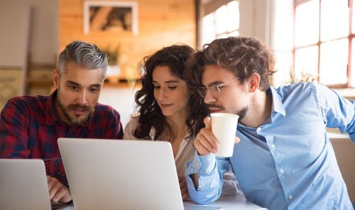 Tres personas jóvenes observando con atención la pantalla de un ordenador portátil en un entorno de oficina o cafetería.