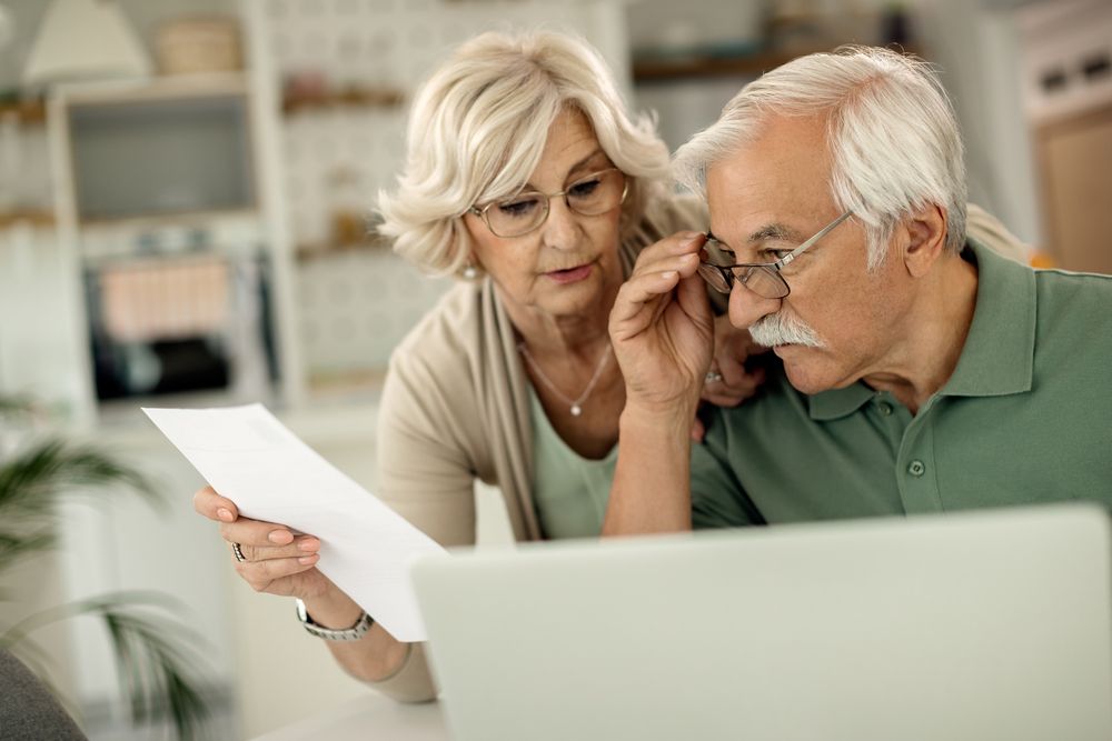 Pareja de personas mayores revisando un documento físico y consultando información en un ordenador portátil.