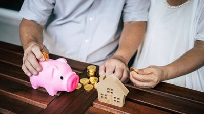 Pareja de jubilados introduciendo monedas en una hucha rosa junto a una pequeña casa de madera.