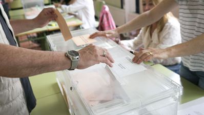 Primer plano de una persona introduciendo un sobre en una urna de plástico transparente durante una jornada electoral.