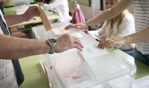 Primer plano de una persona introduciendo un sobre en una urna de plástico transparente durante una jornada electoral.