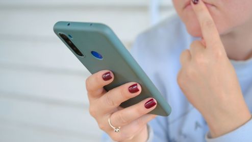 Primer plano de una persona sosteniendo un smartphone con funda verde mientras se lleva un dedo al rostro en actitud pensativa.
