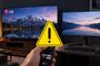 Mano sujetando un mando a distancia frente a un televisor y un monitor LG con un icono de advertencia superpuesto.