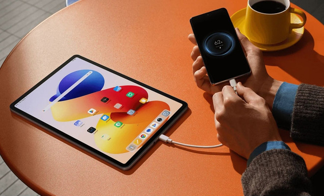 Tableta Redmi Pad Pro conectada mediante un cable USB a un teléfono móvil sobre una mesa naranja.