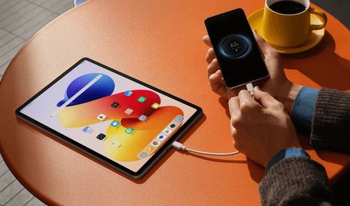 Tableta Redmi Pad Pro conectada mediante un cable USB a un teléfono móvil sobre una mesa naranja.