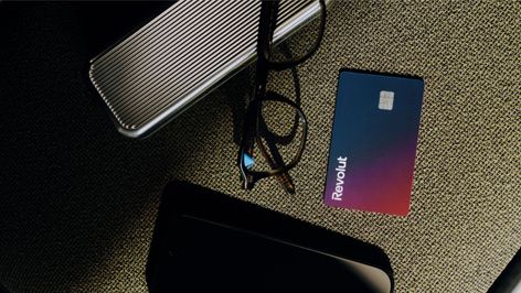 Tarjeta física de Revolut con degradado azul y morado situada junto a unas gafas y objetos personales sobre una superficie de tela.