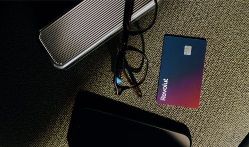 Tarjeta física de Revolut con degradado azul y morado situada junto a unas gafas y objetos personales sobre una superficie de tela.