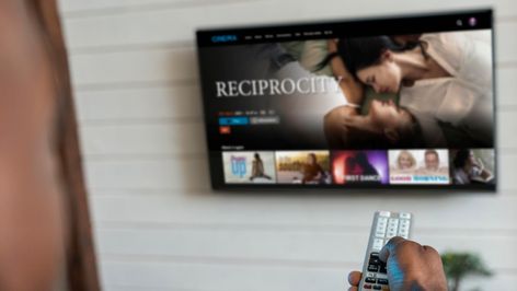 Primer plano de una mano sujetando un mando a distancia frente a una Smart TV con una interfaz de vídeo bajo demanda.