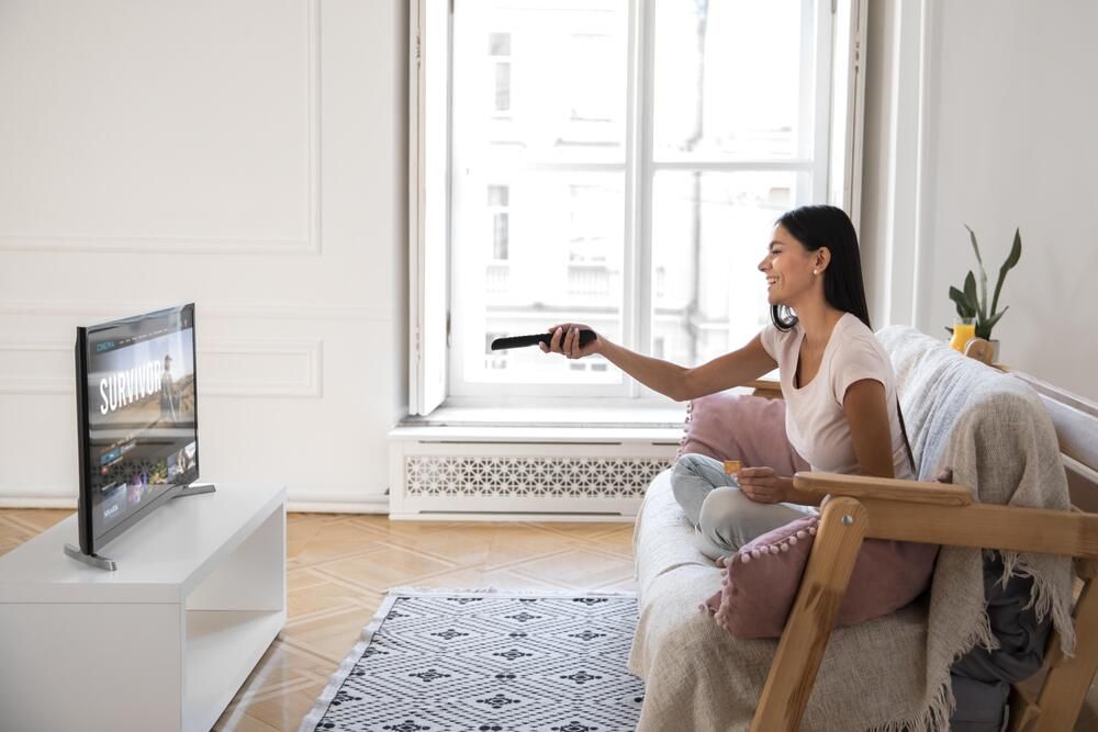 Mujer sentada en un sofá usando el mando a distancia para navegar por el menú de una Smart TV en un salón iluminado.
