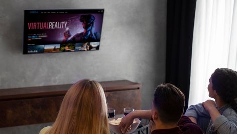 Tres personas de espaldas sentadas frente a una Smart TV colgada en la pared que muestra un menú de realidad virtual.