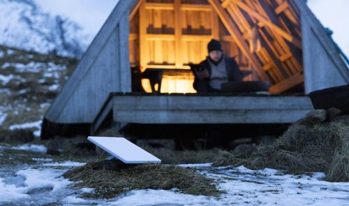 Antena parabólica rectangular de Starlink instalada en un terreno con nieve frente a una cabaña de madera.