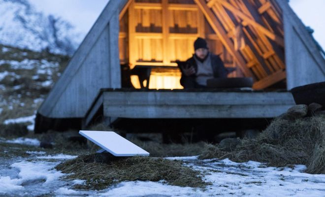 Antena parabólica rectangular de Starlink instalada en un terreno con nieve frente a una cabaña de madera.