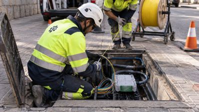 Dos técnicos con chalecos reflectantes y cascos de seguridad trabajan en una arqueta de telecomunicaciones abierta en la acera.