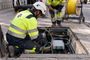 Dos técnicos con chalecos reflectantes y cascos de seguridad trabajan en una arqueta de telecomunicaciones abierta en la acera.