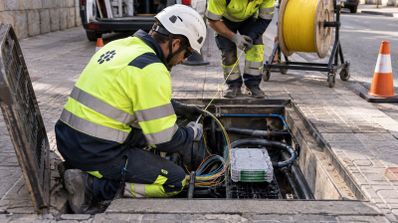 Dos técnicos con chalecos reflectantes y cascos de seguridad trabajan en una arqueta de telecomunicaciones abierta en la acera.