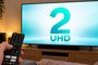Mano sujetando un mando a distancia frente a un televisor que muestra el logotipo de La 2 UHD en pantalla.