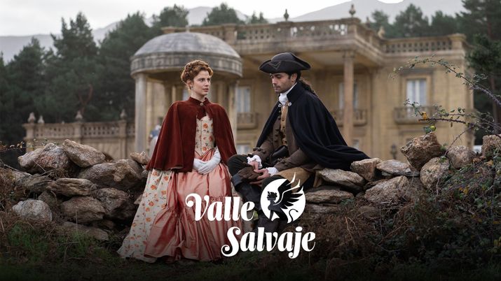 Pareja vestida con trajes de época sentada en un muro de piedra frente a un palacio con el logotipo de Valle Salvaje.