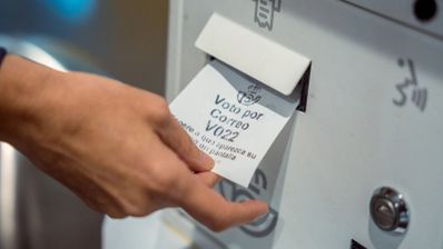 Mano de una persona recogiendo un ticket de turno para el servicio de voto por correo en una oficina postal.