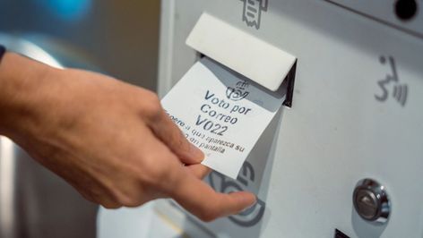 Mano de una persona recogiendo un ticket de turno para el servicio de voto por correo en una oficina postal.
