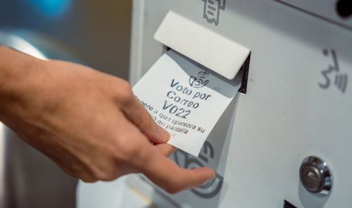 Mano de una persona recogiendo un ticket de turno para el servicio de voto por correo en una oficina postal.